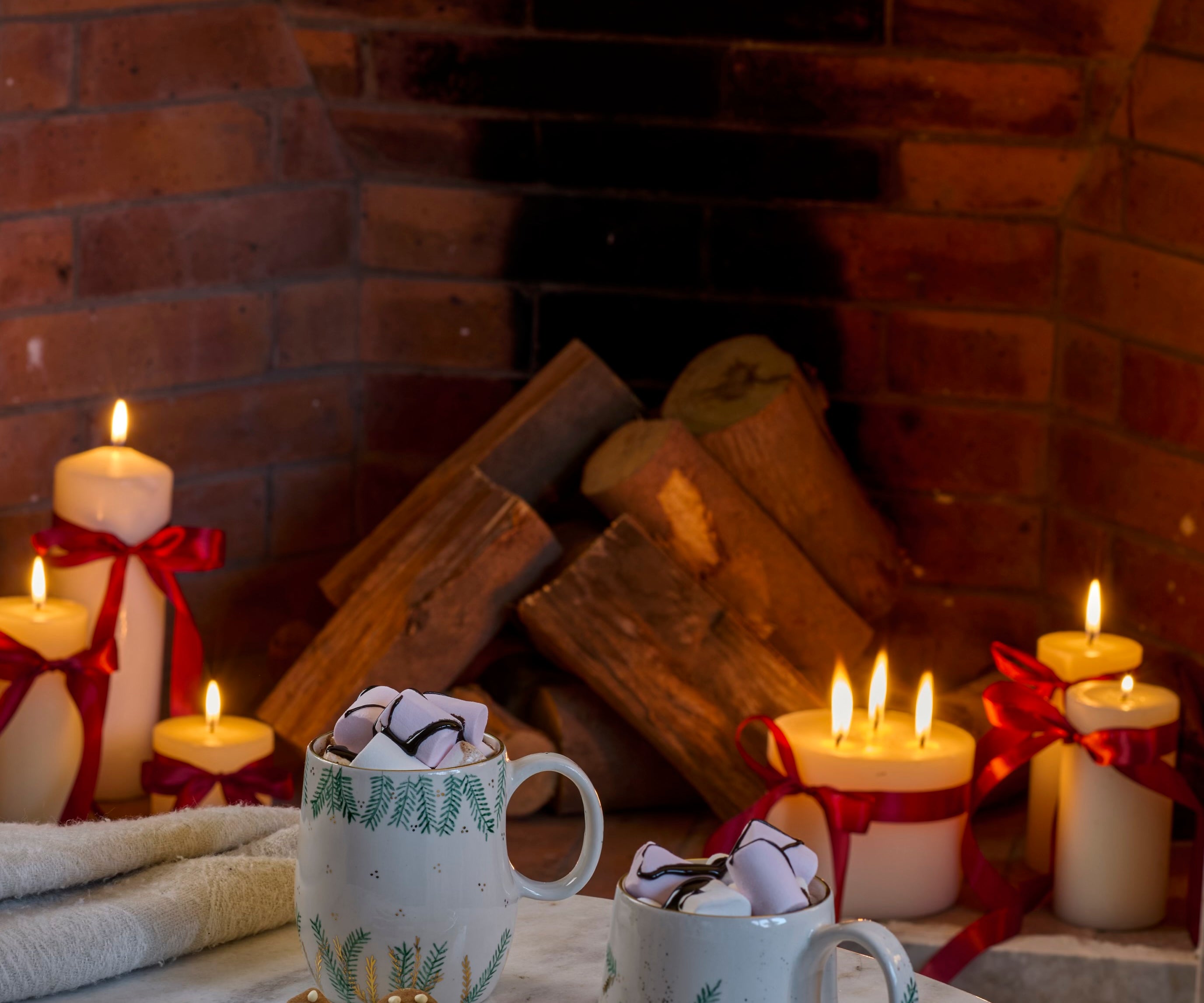 Two mugs with whipped cream and cookies on a marble surface in front of a fireplace with candles.