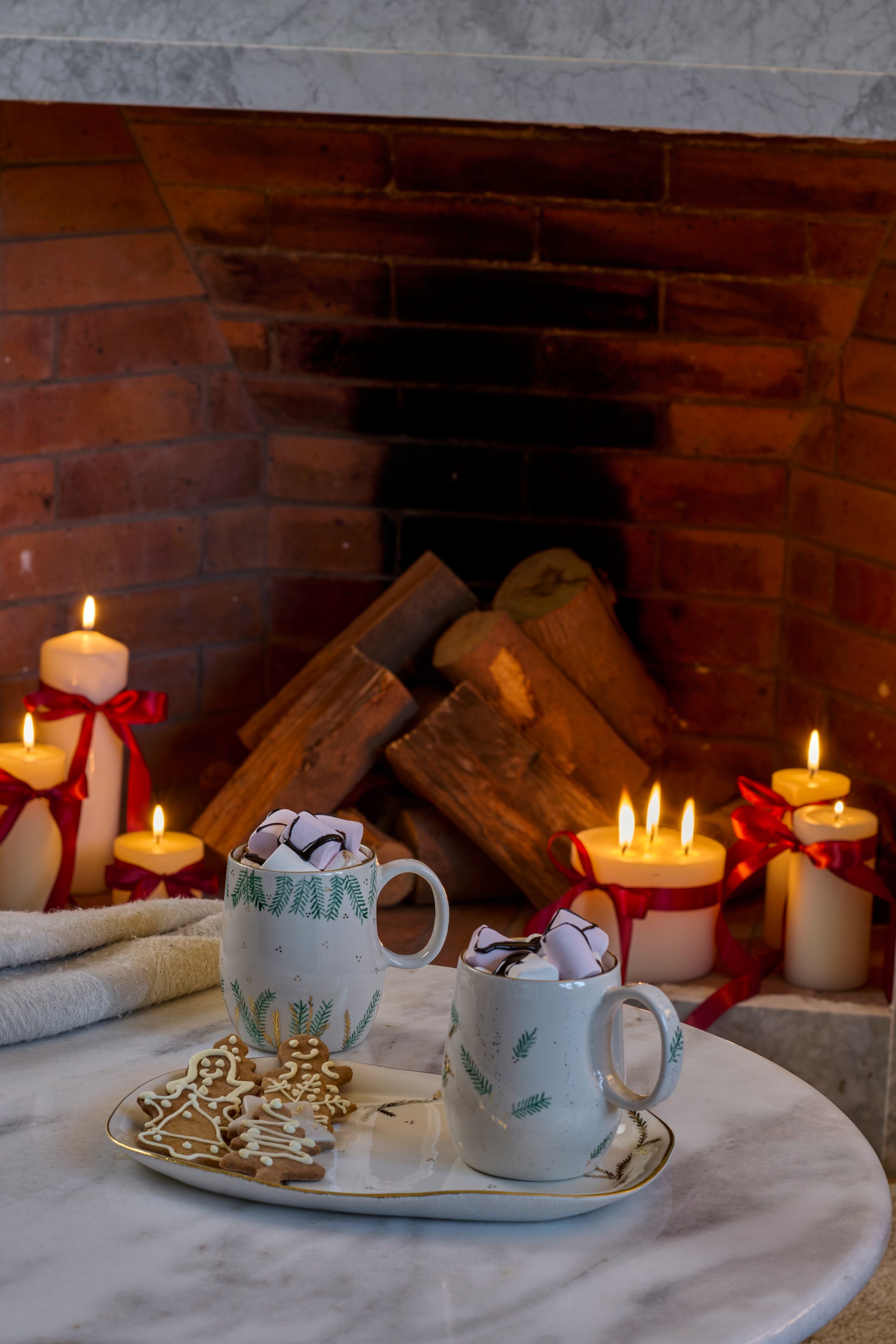 Two mugs with whipped cream and cookies on a marble surface in front of a fireplace with candles.